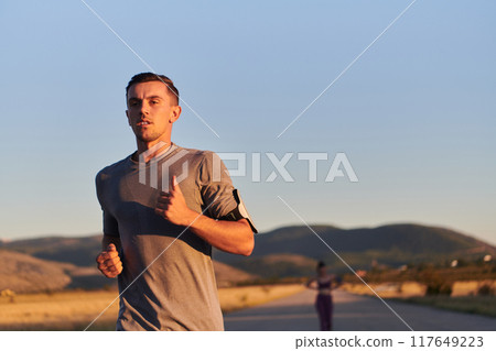 A handsome young couple running together during the early morning hours, with the mesmerizing sunrise casting a warm glow, symbolizing their shared love and vitality A handsome young couple running together during the early morning hours, with the mesmerizing sunrise casting a warm glow, symbolizing their shared love and vitality 117649223