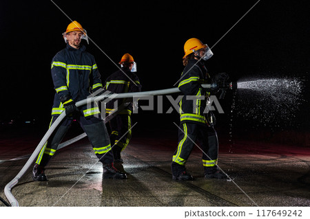 Firefighters using a water hose to eliminate a fire hazard. Team of female and male firemen in dangerous rescue mission. Firefighters using a water hose to eliminate a fire hazard. Team of female and male firemen in dangerous rescue mission. 117649242