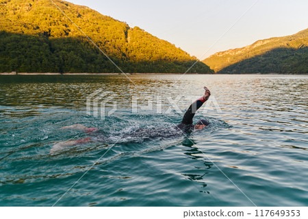 Triathlon athlete swimming on lake in sunrise wearing wetsuit 117649353