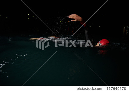 A determined professional triathlete undergoes rigorous night time training in cold waters, showcasing dedication and resilience in preparation for an upcoming triathlon swim competition A determined professional triathlete undergoes rigorous night time training in cold waters, showcasing dedication and resilience in preparation for an upcoming triathlon swim competition 117649378