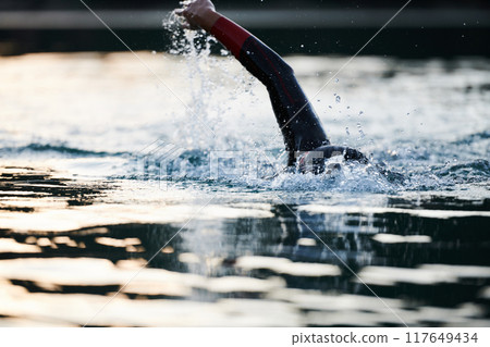 Triathlon athlete swimming on lake in sunrise wearing wetsuit Triathlon athlete swimming on lake in sunrise wearing wetsuit 117649434