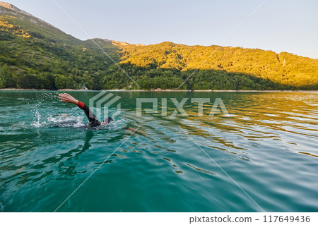 Triathlon athlete swimming on lake in sunrise wearing wetsuit Triathlon athlete swimming on lake in sunrise wearing wetsuit 117649436