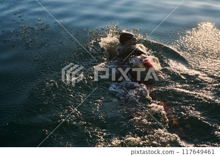Triathlon athlete swimming on lake in sunrise wearing wetsuit 117649461