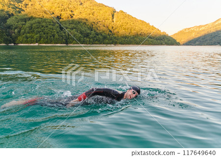 Triathlon athlete swimming on lake in sunrise wearing wetsuit Triathlon athlete swimming on lake in sunrise wearing wetsuit 117649640