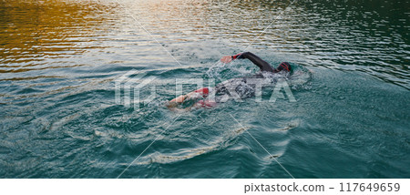 A professional triathlete trains with unwavering dedication for an upcoming competition at a lake, emanating a sense of athleticism and profound commitment to excellence. 117649659