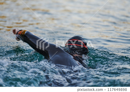 Triathlon athlete swimming on lake in sunrise wearing wetsuit 117649898