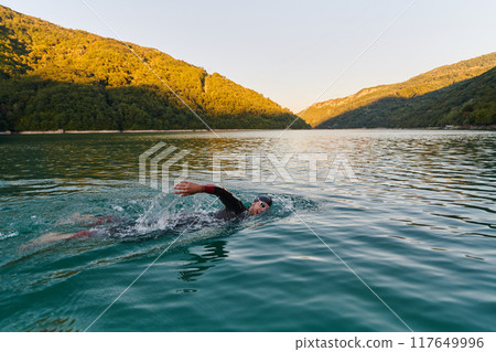 Triathlon athlete swimming on lake in sunrise wearing wetsuit Triathlon athlete swimming on lake in sunrise wearing wetsuit 117649996
