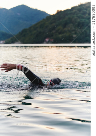 A professional triathlete trains with unwavering dedication for an upcoming competition at a lake, emanating a sense of athleticism and profound commitment to excellence. 117650092