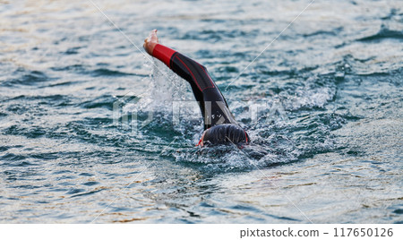 A professional triathlete trains with unwavering dedication for an upcoming competition at a lake, emanating a sense of athleticism and profound commitment to excellence. 117650126