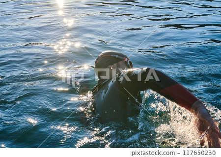 Triathlon athlete swimming on lake in sunrise wearing wetsuit Triathlon athlete swimming on lake in sunrise wearing wetsuit 117650230