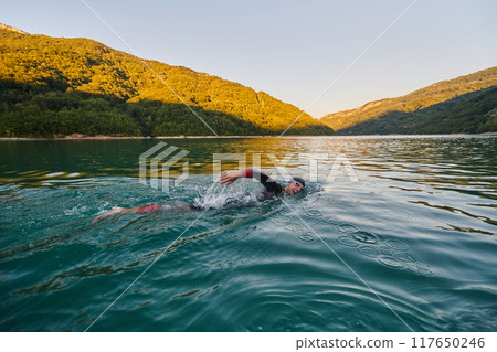 Triathlon athlete swimming on lake in sunrise wearing wetsuit Triathlon athlete swimming on lake in sunrise wearing wetsuit 117650246
