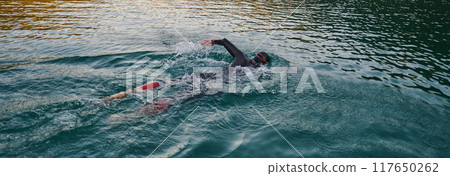 A professional triathlete trains with unwavering dedication for an upcoming competition at a lake, emanating a sense of athleticism and profound commitment to excellence. 117650262