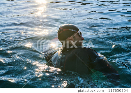 Triathlon athlete swimming on lake in sunrise wearing wetsuit 117650304