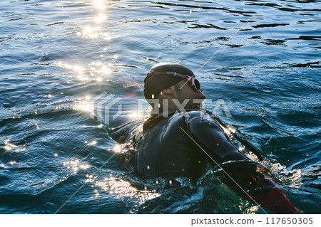 Triathlon athlete swimming on lake in sunrise wearing wetsuit 117650305