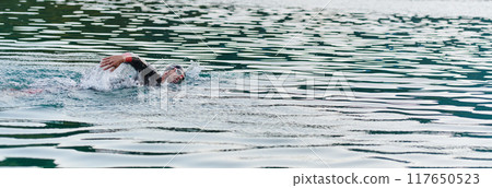 A professional triathlete trains with unwavering dedication for an upcoming competition at a lake, emanating a sense of athleticism and profound commitment to excellence. 117650523