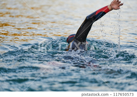 Triathlon athlete swimming on lake in sunrise wearing wetsuit Triathlon athlete swimming on lake in sunrise wearing wetsuit 117650573