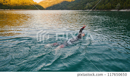 Triathlon athlete swimming on lake in sunrise wearing wetsuit Triathlon athlete swimming on lake in sunrise wearing wetsuit 117650650