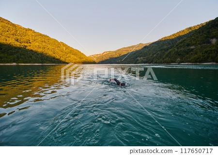 Triathlon athlete swimming on lake in sunrise wearing wetsuit Triathlon athlete swimming on lake in sunrise wearing wetsuit 117650717