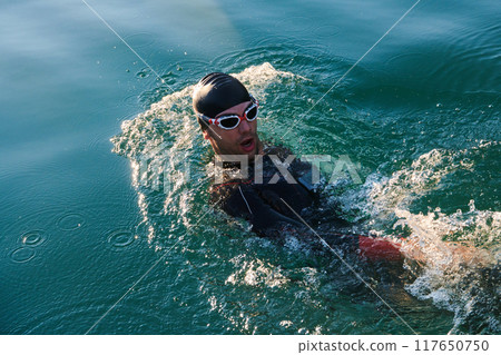 Triathlon athlete swimming on lake in sunrise wearing wetsuit 117650750