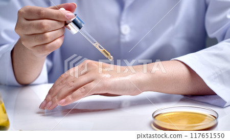 Close-up photo from the front of a laboratory scene, a scientist testing cosmetics on the back of his hand, next to a glass petri dish contains yellow liquid. Photo for advertising, space for text Close-up photo from the front of a laboratory scene, a scientist testing cosmetics on the back of his hand, next to a glass petri dish contains yellow liquid. Photo for advertising, space for text 117651550