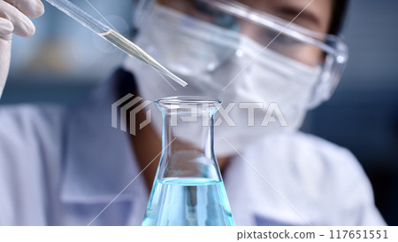 Macro front angle view of a scientist is dropping light blue liquid substance into an erlenmeyer flask. Concept of substance study experiment on blurred lab background, for promotional photo Macro front angle view of a scientist is dropping light blue liquid substance into an erlenmeyer flask. Concept of substance study experiment on blurred lab background, for promotional photo 117651551