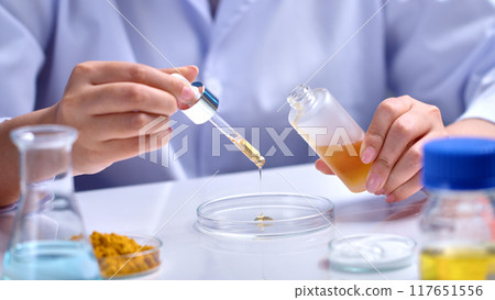 Cosmetic product experiment in science lab scene from front view. A serum bottle being held by a scientist, yellow liquid dripped into a glass petri dish, which placed on table with several lab items Cosmetic product experiment in science lab scene from front view. A serum bottle being held by a scientist, yellow liquid dripped into a glass petri dish, which placed on table with several lab items 117651556