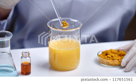 Advertising photo for turmeric cosmetic product in lab scene, was taken from close up front view. The glass spoon of turmeric powder prepare to put into glass beaker, which placed on white table 117651561