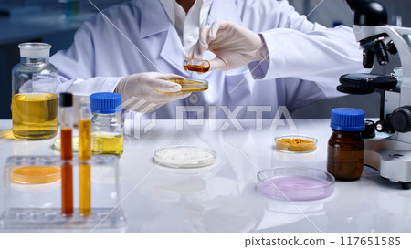 The researcher pours yellow liquid from an experiment sample storage vial into glass petri dish. White table displays many lab items like petri dish, chemical bottle, microscope and test tube The researcher pours yellow liquid from an experiment sample storage vial into glass petri dish. White table displays many lab items like petri dish, chemical bottle, microscope and test tube 117651585