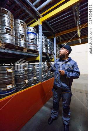 Brewery technologist inspecting rows of kegs in the storage area, ensuring they are properly labeled and ready for distribution. Maintaining accuracy in inventory tracking. Brewery technologist inspecting rows of kegs in the storage area, ensuring they are properly labeled and ready for distribution. Maintaining accuracy in inventory tracking. 117654451
