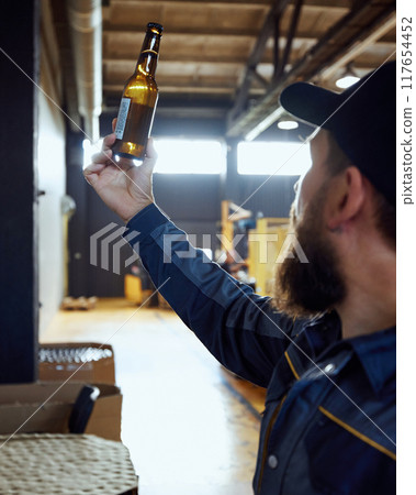 Focused man, brewery technologist examining beer bottle in the packaging area, ensuring consistency and quality. Focused man, brewery technologist examining beer bottle in the packaging area, ensuring consistency and quality. 117654452