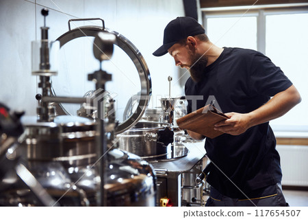 Bearded man, technologist checking brewing tools, making notes, demonstrating expertise needed to crafting beer. 117654507