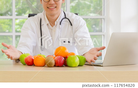 Male doctor in white coat with stethoscope at office desk with laptop and presenting variety of fresh fruits including apples, oranges and kiwis. 117654820