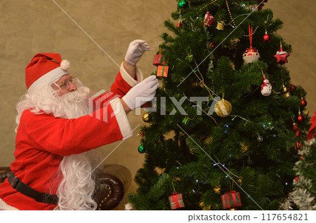 Santa Claus sitting on leather sofa and reaching up to hang ornaments on decorates Christmas tree at home. 117654821
