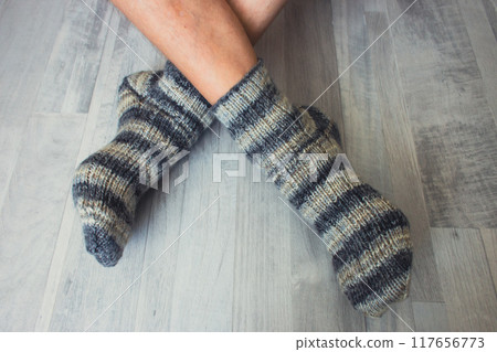 Men legs in knitted striped socks. Woolen knitted socks on crossed legs. Autumn warm clothing. Striped crochet pattern for socks. Handmade fashion. Cozy lifestyle. 117656773