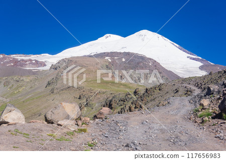 highlands landscape, road through a deserted rocky high mountain plateau and the double top of Mount Elbrus in the distance highlands landscape, road through a deserted rocky high mountain plateau and the double top of Mount Elbrus in the distance 117656983