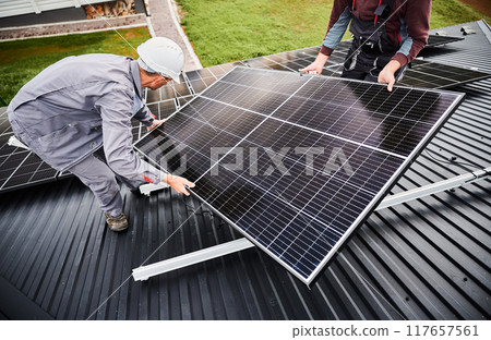 Technicians installing solar panel system on roof of house. Men Installers in helmets carrying photovoltaic solar module outdoors. Concept of alternative and renewable energy. 117657561