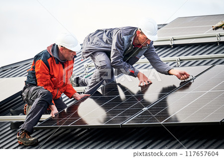 Roofers installing photovoltaic solar panels on roof of house. Men engineers in helmets building solar module system with help of hex key. Concept of alternative, renewable energy. Roofers installing photovoltaic solar panels on roof of house. Men engineers in helmets building solar module system with help of hex key. Concept of alternative, renewable energy. 117657604