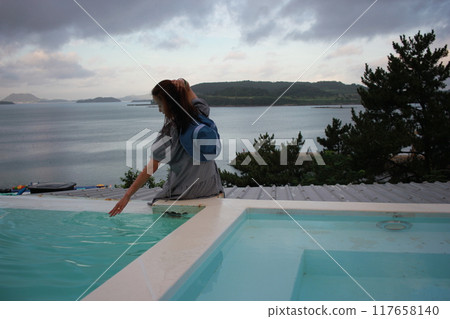Dolsan-eup, Yeosu-si, South Korea - July 26, 2024: Young woman watching sunrise at a resort with a swimming pool overlooking the sea in Yeosu Dolsan-eup, Yeosu-si, South Korea - July 26, 2024: Young woman watching sunrise at a resort with a swimming pool overlooking the sea in Yeosu 117658140