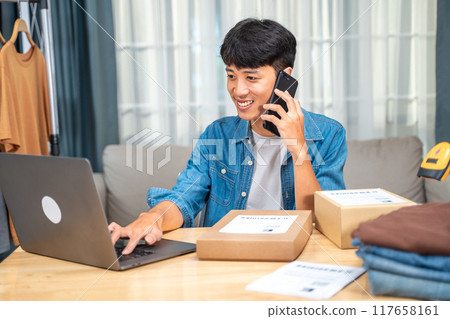 Happy young online seller man talking to his customer on the phone while checking incoming orders from his laptop computer Happy young online seller man talking to his customer on the phone while checking incoming orders from his laptop computer 117658161