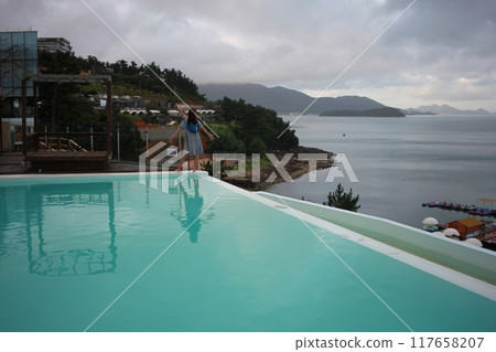 Dolsan-eup, Yeosu-si, South Korea - July 26, 2024: Young woman watching sunrise at a resort with a swimming pool overlooking the sea in Yeosu Dolsan-eup, Yeosu-si, South Korea - July 26, 2024: Young woman watching sunrise at a resort with a swimming pool overlooking the sea in Yeosu 117658207