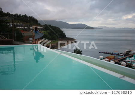 Dolsan-eup, Yeosu-si, South Korea - July 26, 2024: Young woman watching sunrise at a resort with a swimming pool overlooking the sea in Yeosu Dolsan-eup, Yeosu-si, South Korea - July 26, 2024: Young woman watching sunrise at a resort with a swimming pool overlooking the sea in Yeosu 117658210