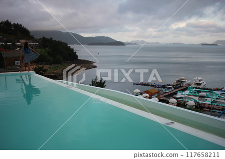 Dolsan-eup, Yeosu-si, South Korea - July 26, 2024: Young woman watching sunrise at a resort with a swimming pool overlooking the sea in Yeosu Dolsan-eup, Yeosu-si, South Korea - July 26, 2024: Young woman watching sunrise at a resort with a swimming pool overlooking the sea in Yeosu 117658211