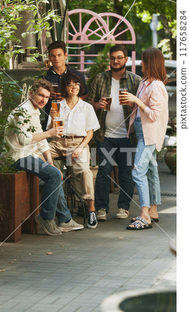 Relaxed and happy young people, men and women, friends gathering at cozy outdoor bar, pub, enjoying regressing beer. Festive atmosphere Relaxed and happy young people, men and women, friends gathering at cozy outdoor bar, pub, enjoying regressing beer. Festive atmosphere 117658284
