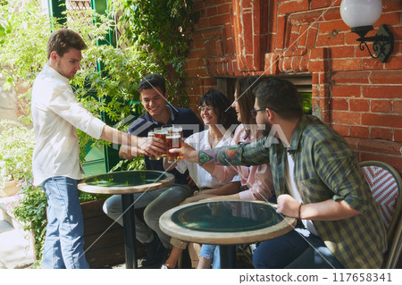 Lively group of young people, men and women, friends gathering at local outdoor bar, clinking beer glasses, enjoying good company Lively group of young people, men and women, friends gathering at local outdoor bar, clinking beer glasses, enjoying good company 117658341