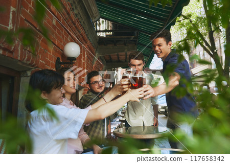 Smiling happy young people, men and women, friends gathering at outdoor bar in warm sunny evening and enjoying refreshing beer. Cheers. Smiling happy young people, men and women, friends gathering at outdoor bar in warm sunny evening and enjoying refreshing beer. Cheers. 117658342
