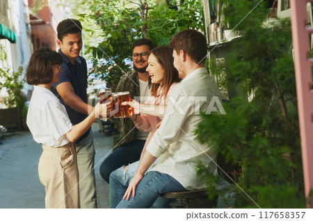 Lively group of friends, young people meeting at outdoor modern bar, clinking beer glasses and having good relaxing time on warm day 117658357