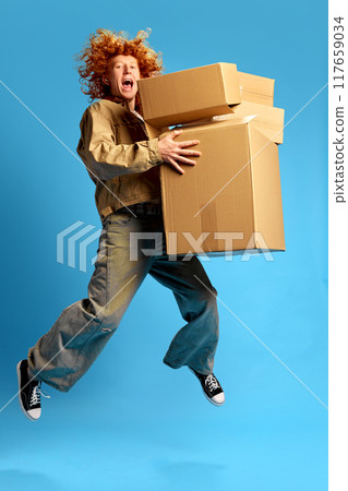 Overjoyed young red haired man, shopaholic, jumping holding cardboard boxes against blue studio background. Concept of Black Friday sale. 117659034