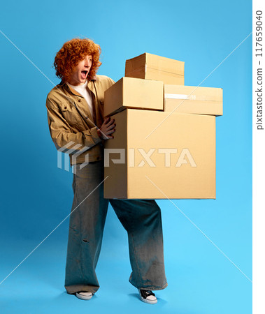 Surprised young man dressed stylish attire holding heavy, huge stack of delivery boxes against blue studio background. Concept of Black Friday sale. Surprised young man dressed stylish attire holding heavy, huge stack of delivery boxes against blue studio background. Concept of Black Friday sale. 117659040