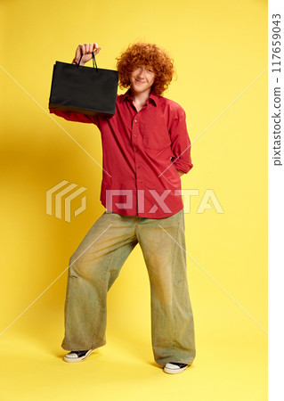 Delightful young man dressed vintage stylish outfit holding black back with purchases against yellow background. Concept of Black Friday sale. 117659043