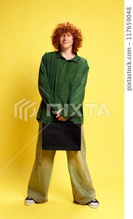 Full length photo of young man in retro style clothes holding black package against yellow background. Concept of Black Friday sale. 117659048